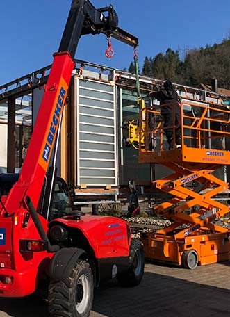 Ein roter Kran hebt Baumaterial für eine Montagearbeit, dahinter eine orangefarbene Hebebühne.