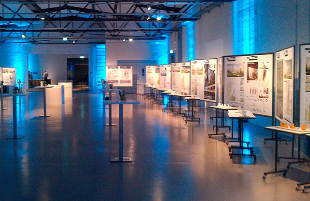 Ausstellungshalle eines Architekturwettbewerbs: weiße Pinnwände sind mit Postern bestückt, davor stehen Architekturmodelle auf Modelltischen.