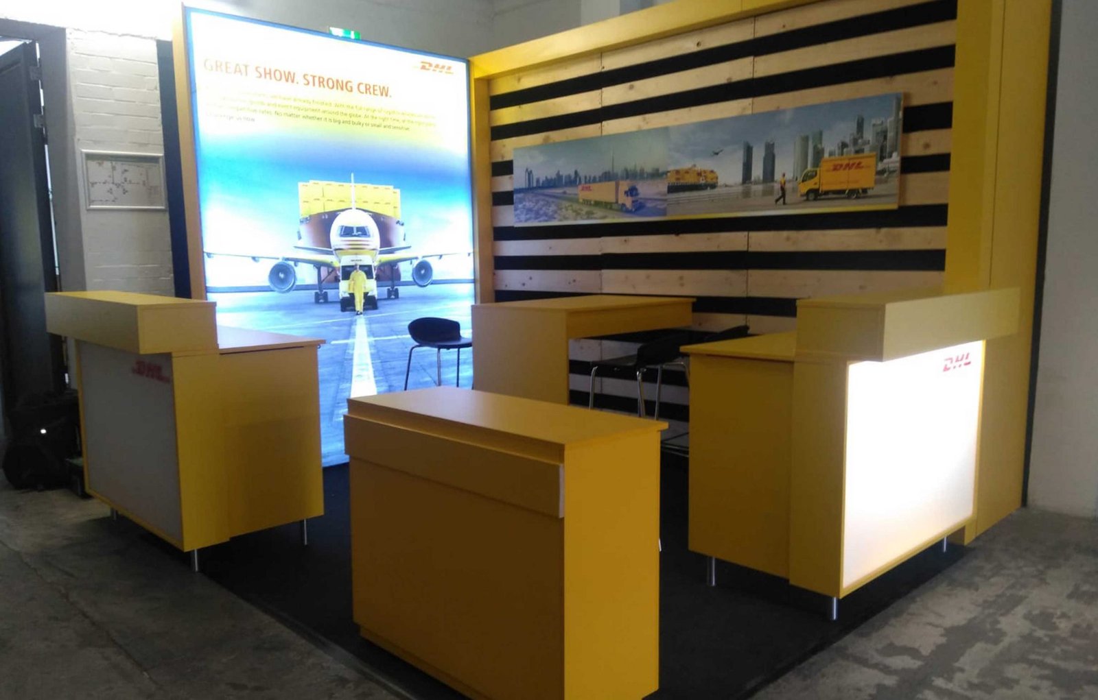 DHL Messestand mit gelben Bauelementen: Tresen, Regale, eine Leuchtwand.